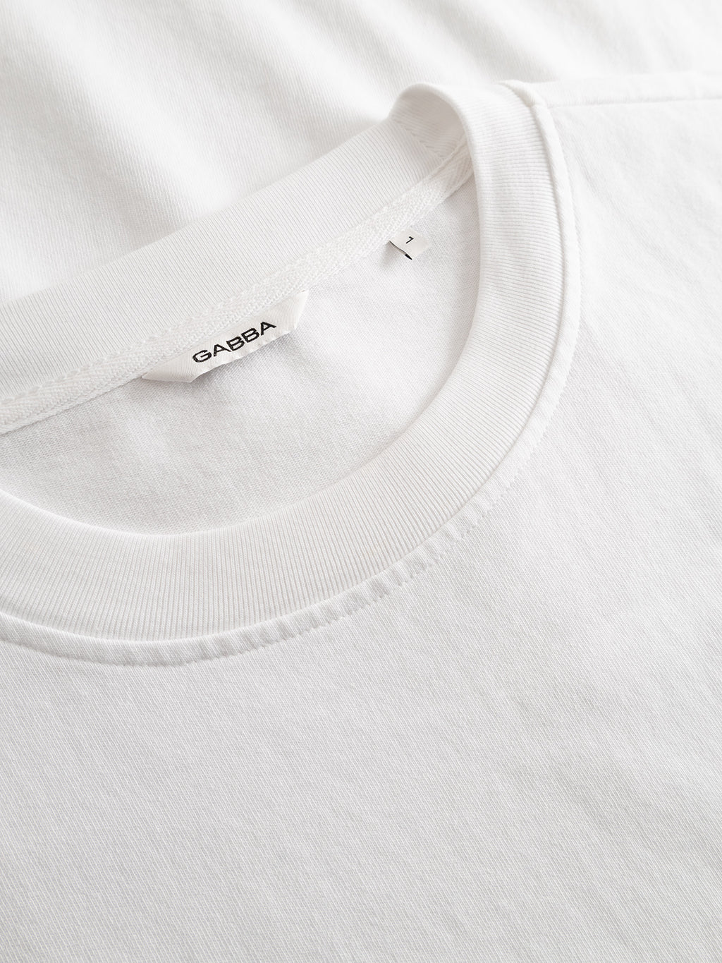 A close-up of the Gabba GABnigel Wix Pkt Boxy Hour SS T-Shirt in white, a plain heavyweight cotton crew neck featuring a visible Gabba label inside the collar, laid on top of another white shirt.