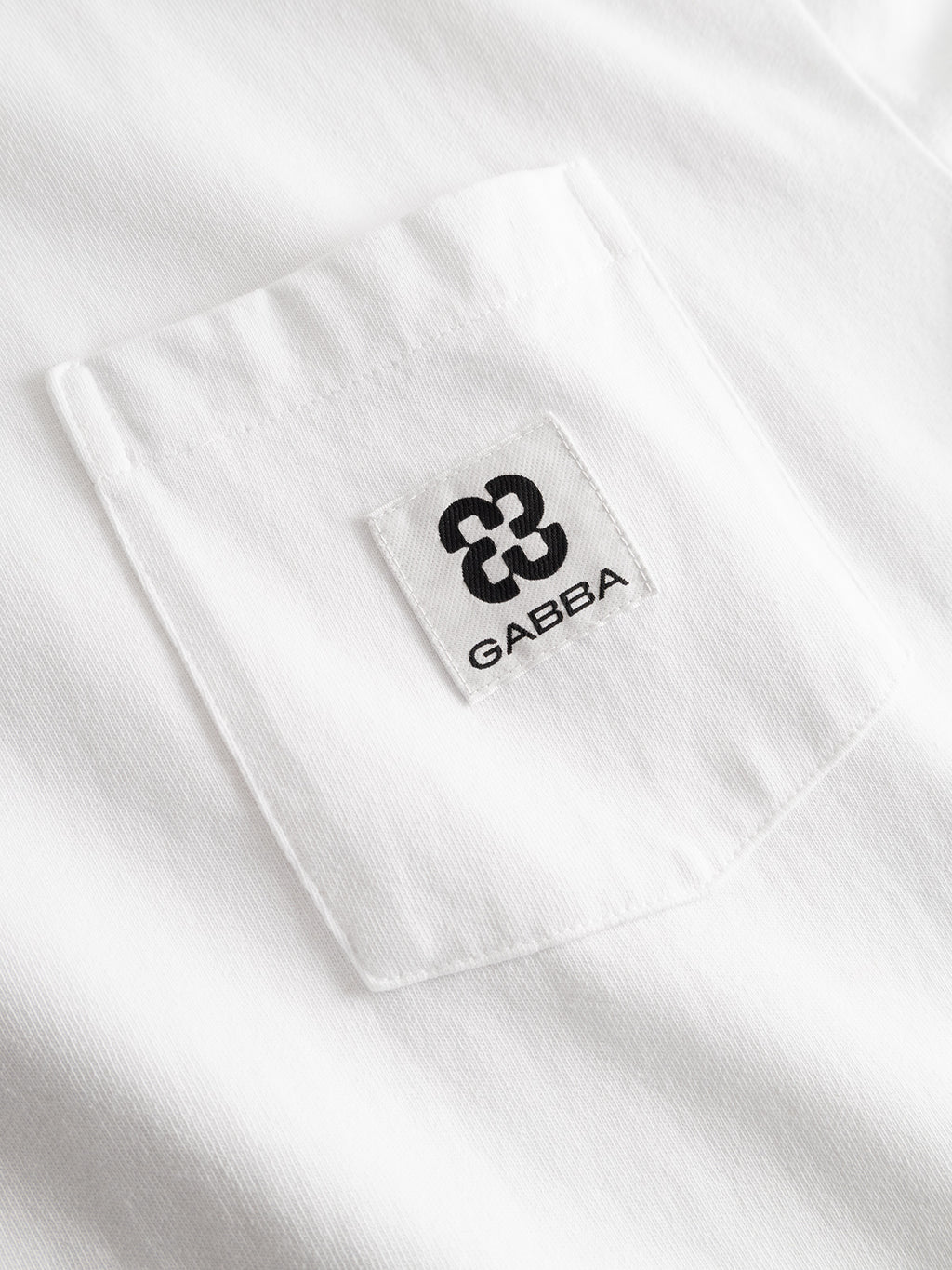 Close-up of a white GABnigel Wix Pkt Boxy Hour SS T-Shirt by Gabba, crafted from heavyweight cotton. The expertly stitched chest pocket features a small white label with a black abstract logo and the brand name Gabba.