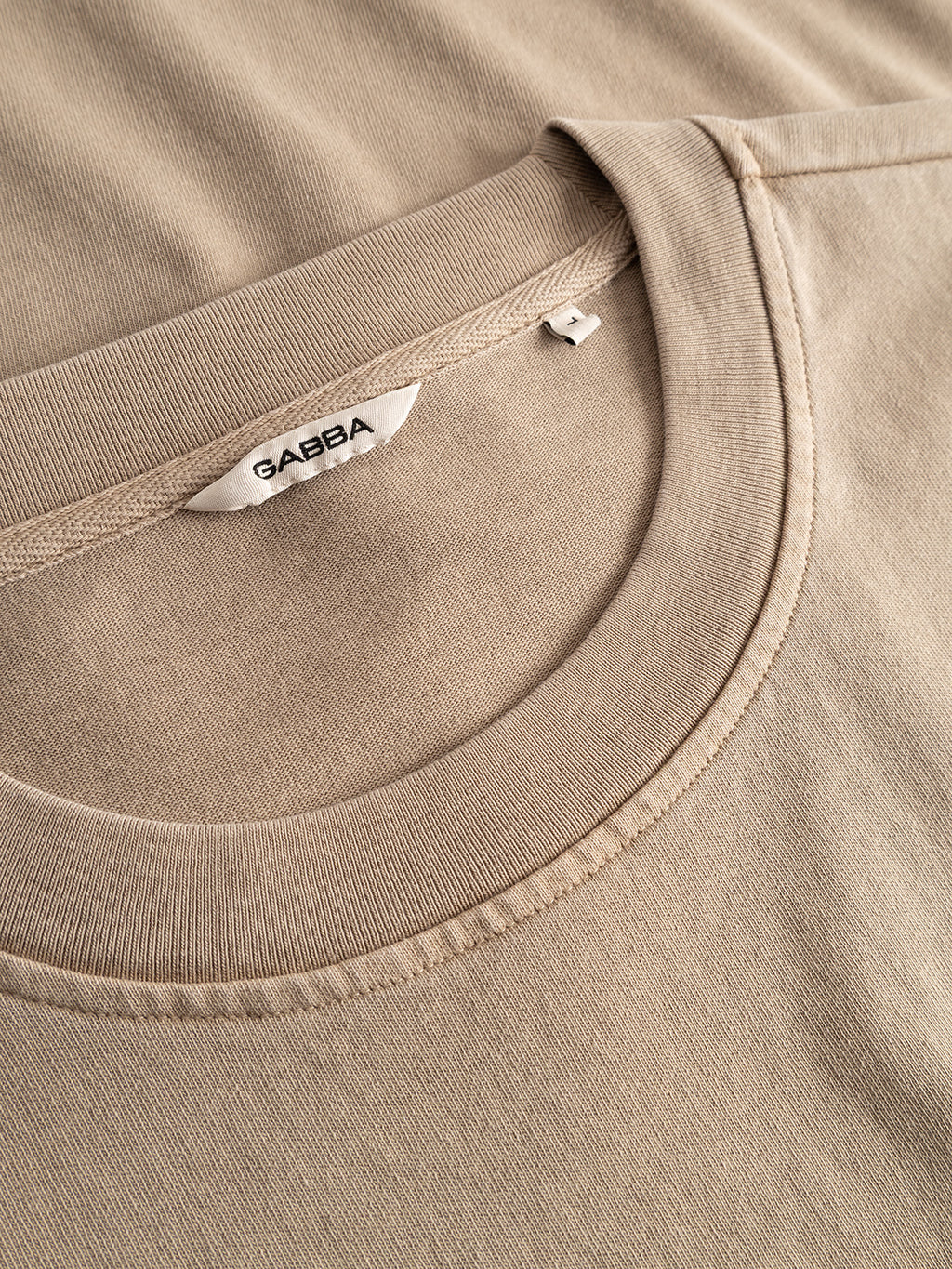 The GABnigel Wix Pkt Boxy Hour SS T-Shirt in Sand Crocker by Gabba is shown laying flat, highlighting its round neckline and a white tag with the brand’s name. Crafted from heavyweight cotton, the fabric texture and stitching details are visible.