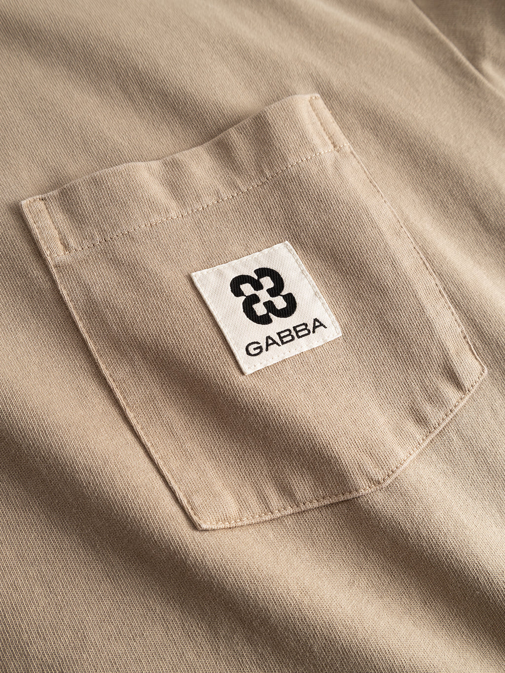 A close-up of the GABnigel Wix Pkt Boxy Hour SS T-Shirt in Sand Crocker, showcasing its beige heavyweight cotton fabric pocket with a white label featuring Gabba’s black abstract logo and branding.