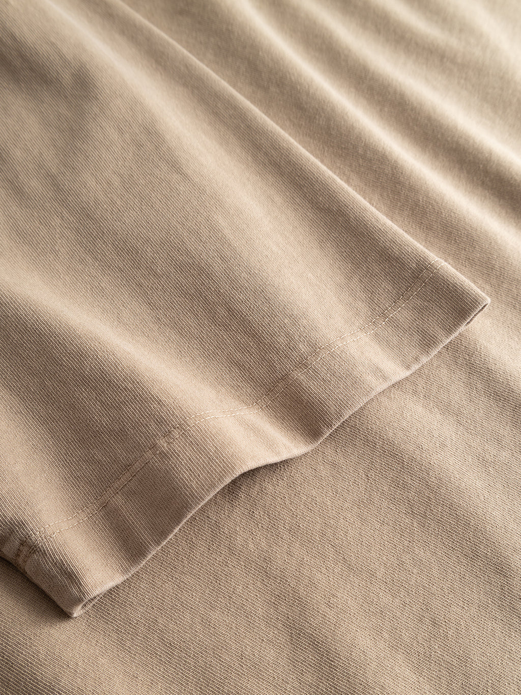 A close-up of the Gabba GABnigel Wix Pkt Boxy Hour SS T-Shirt in Sand Crocker showcases heavyweight cotton fabric in a beige hue, with detailed hem and stitching visible. The soft texture, subtle shadows, and folds highlight the boxy t-shirt design.