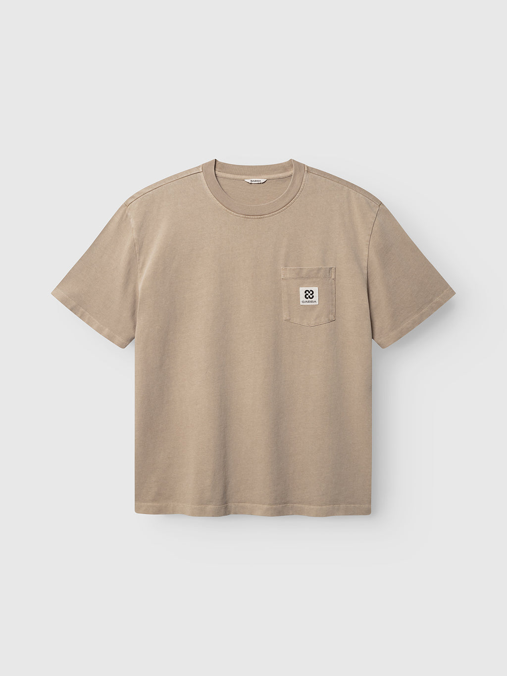 Introducing the Gabba GABnigel Wix Pkt Boxy Hour SS T-Shirt in Sand Crocker. This beige, short-sleeve t-shirt features a boxy silhouette with a classic crew neckline and a chest pocket. Made from heavyweight cotton, the pocket is adorned with a subtle white and black geometric logo patch. The shirt is presented against a plain white background.