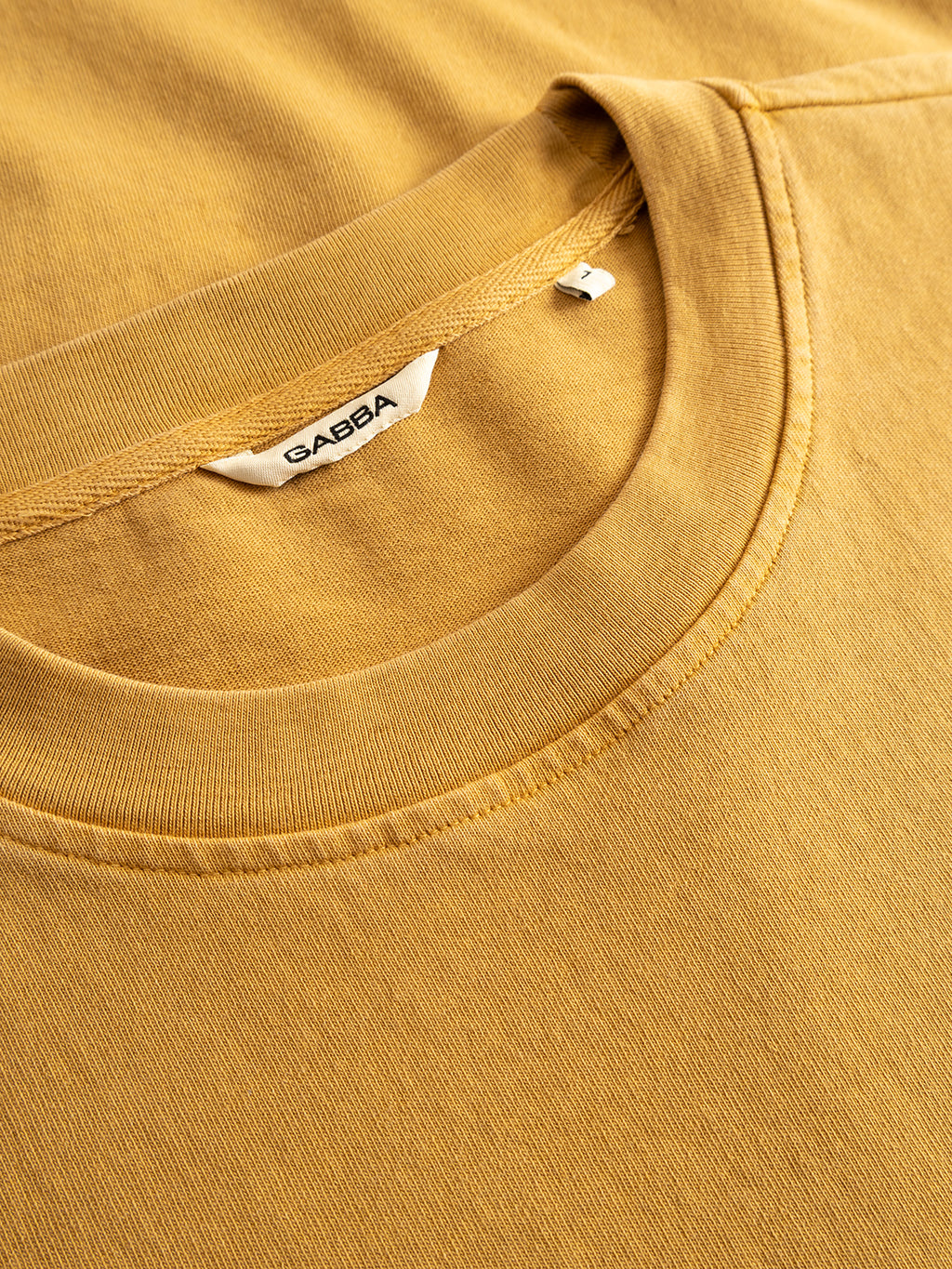 A close-up of the Gabba GABnigel Wix Pkt Boxy Hour SS T-Shirt in mustard yellow, featuring a boxy silhouette, visible stitching, and a white brand label inside the collar, crafted from soft heavyweight cotton.