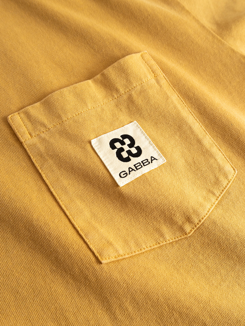 Close-up of the GABnigel Wix Pkt Boxy Hour SS T-Shirt in Mustard Yellow by Gabba, showcasing its heavyweight cotton pocket with a white label featuring the signature Gabba Brand Mark—an abstract black logo above the word GABBA. The boxy silhouette enhances its modern appeal.
