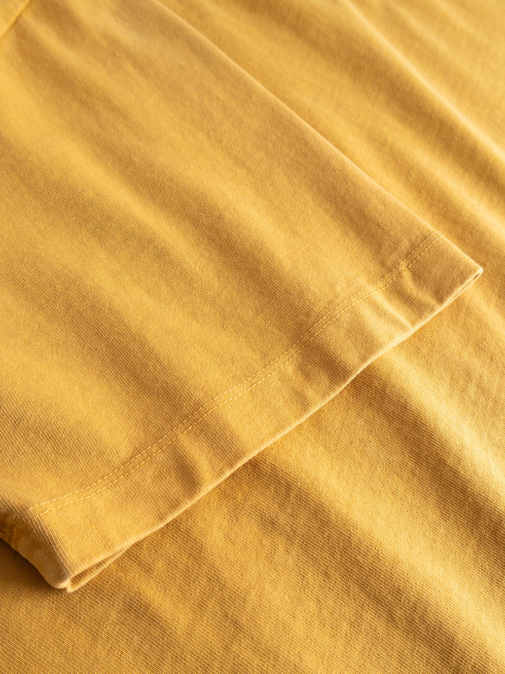 A close-up of the GABnigel Wix Pkt Boxy Hour SS T-Shirt in Mustard Yellow by Gabba shows the boxy sleeve lying flat on matching fabric, highlighting the shirt’s hem, precise stitching, and the rich texture of heavyweight cotton. The subtle Gabba Brand Mark adds a distinctive touch.