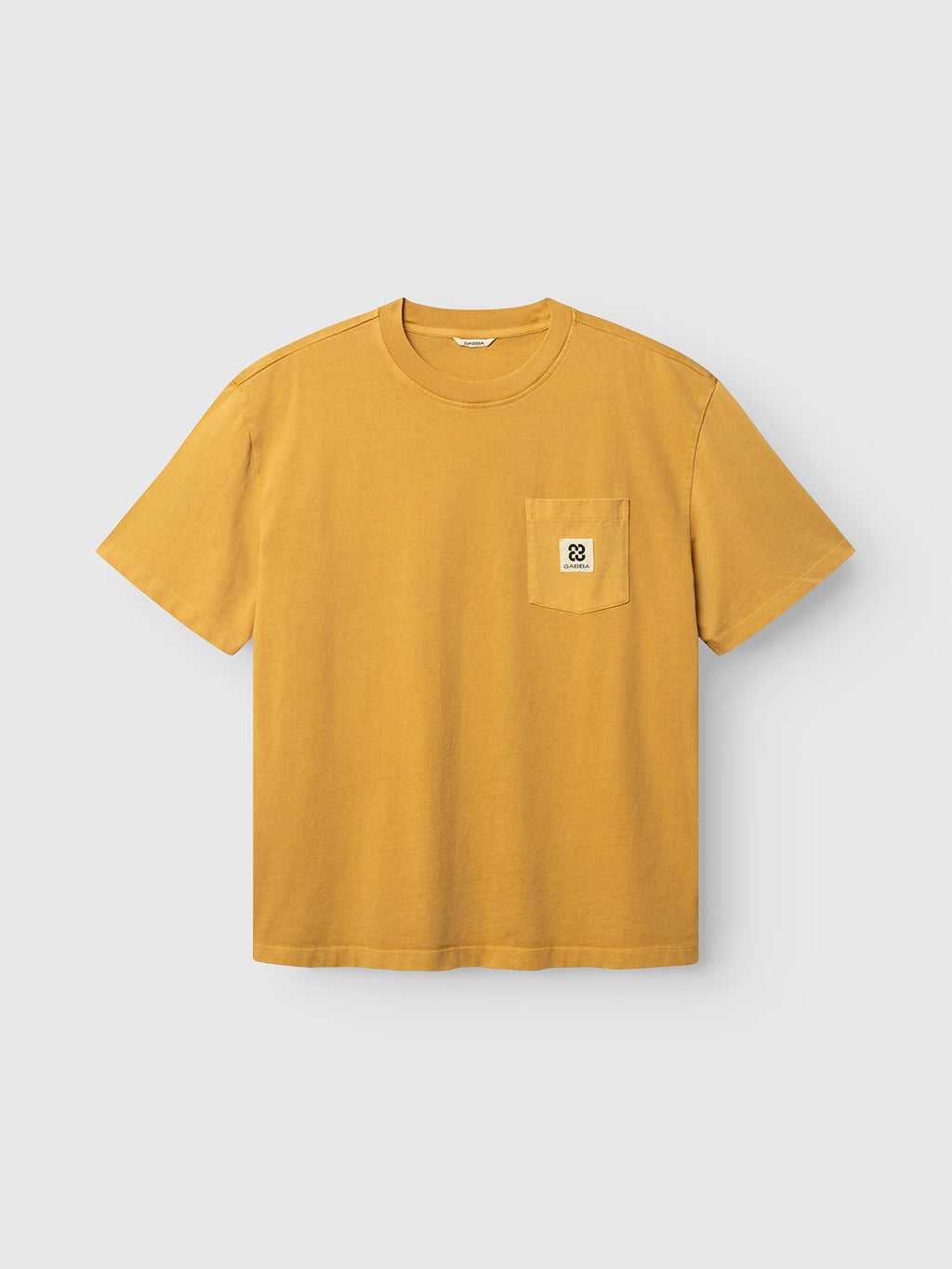 The GABnigel Wix Pkt Boxy Hour SS T-Shirt by Gabba is a mustard yellow heavyweight cotton short-sleeve tee, featuring a boxy fit, round neck, and a small left chest pocket adorned with a white patch displaying a black minimalist design and the Gabba Brand Mark. The t-shirt is photographed laid flat on a plain light background.