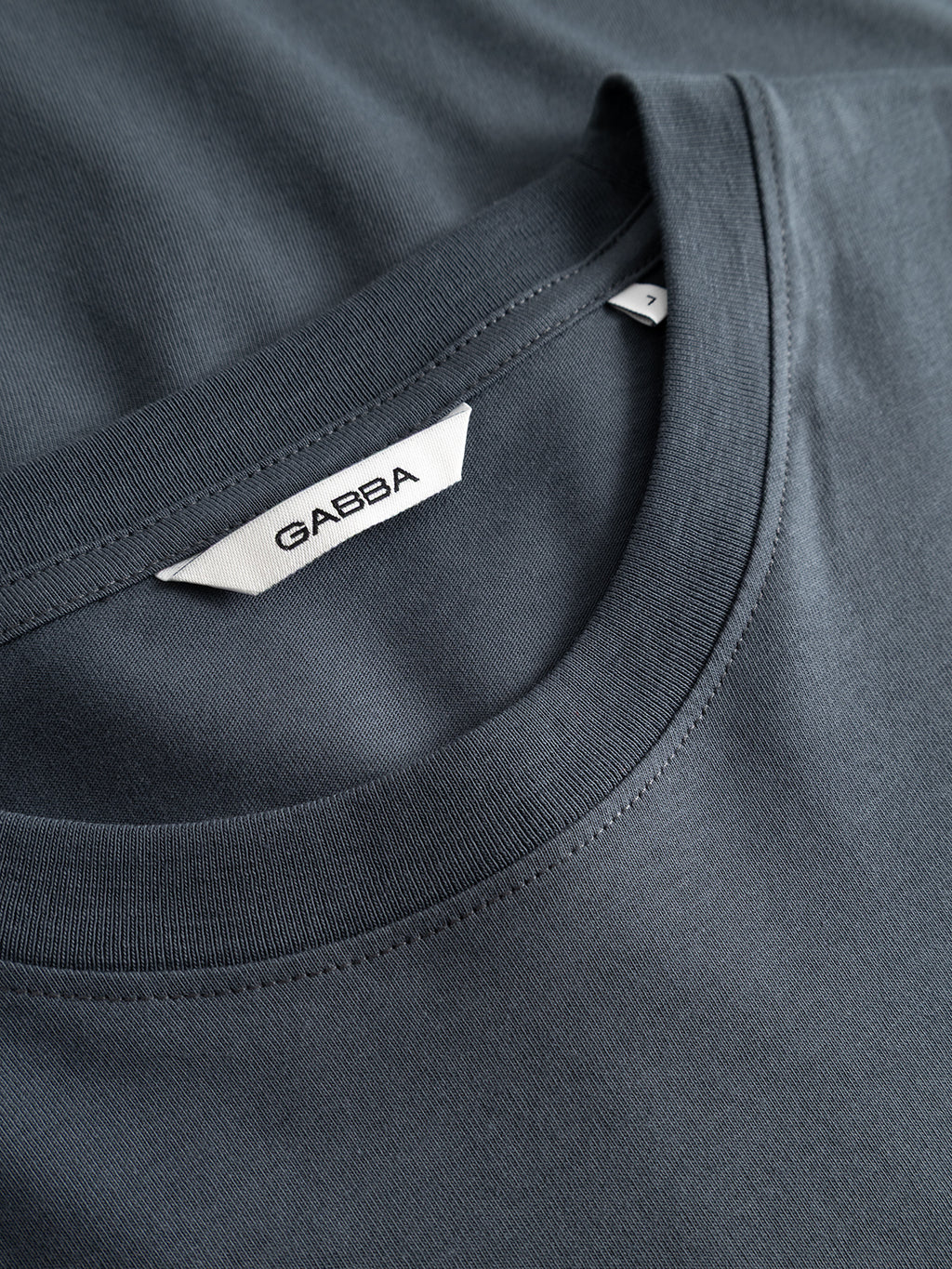 Close-up of the GABdune Element GOTS SS T-Shirt in Turbulence Blue by Gabba, featuring a white Gabba label sewn inside the collar. The organic cotton fabric appears smooth and soft, highlighting its premium GOTS-certified quality as it’s laid out atop another similar garment.