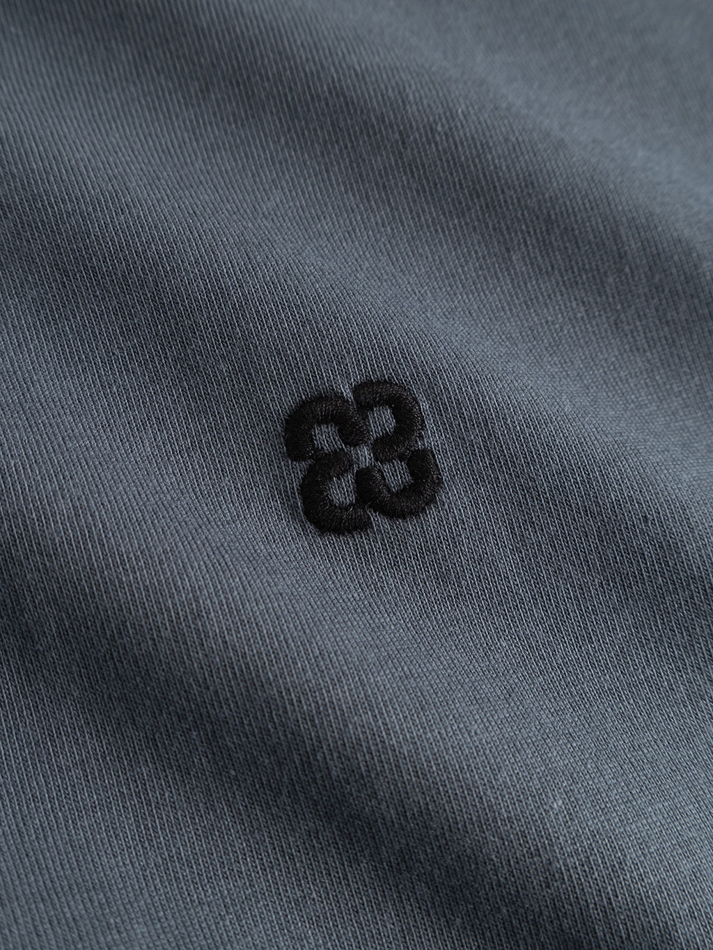 A close-up of the turbulence blue fabric from the Gabba GABdune Element GOTS SS T-Shirt, made from GOTS-certified organic cotton, featuring a small black abstract embroidered logo with four interlocking shapes at the center.