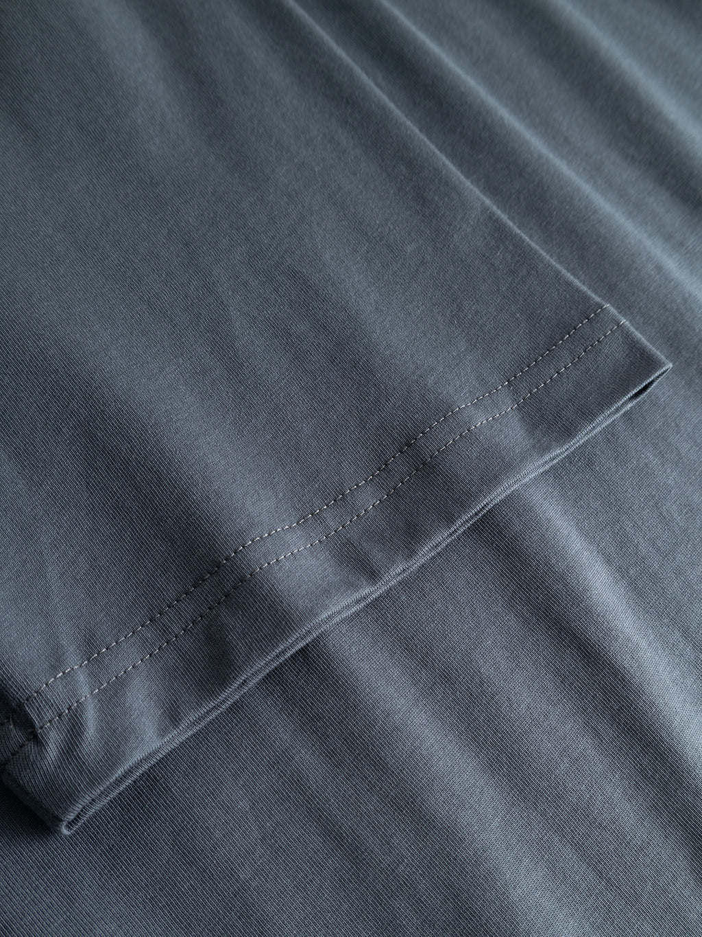 Close-up of the hem and stitching on a Gabba GABdune Element GOTS SS T-Shirt in Turbulence Blue, possibly at the sleeve, laid flat on a matching surface. The visible stitching runs parallel to the edge of the GOTS-certified organic cotton fabric.
