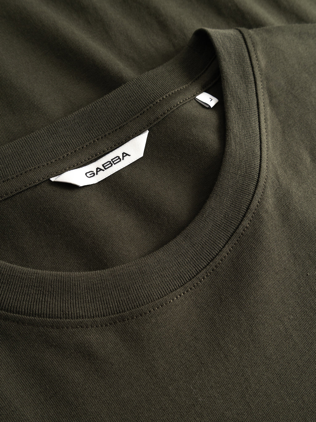 Close-up of the GABdune Element GOTS SS T-Shirt in Forest Night, an organic cotton t-shirt by Gabba. The soft, smooth dark green fabric features a white neck label displaying the Gabba brand name, with another shirt in the same color partially visible underneath.