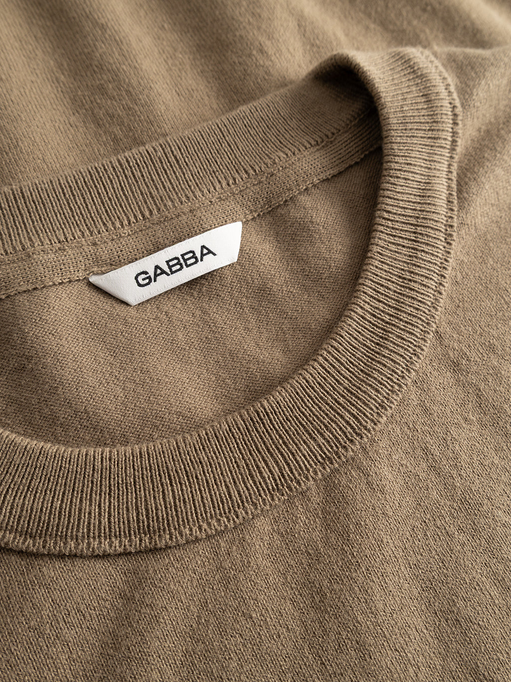 Close-up of the Gabba GABfumo Element Spy LS Knit in Sand Crocker—a beige crew neck sweater featuring a white GABBA label sewn inside the collar. The flat-knit cotton fabric looks soft, with neatly stitched details.