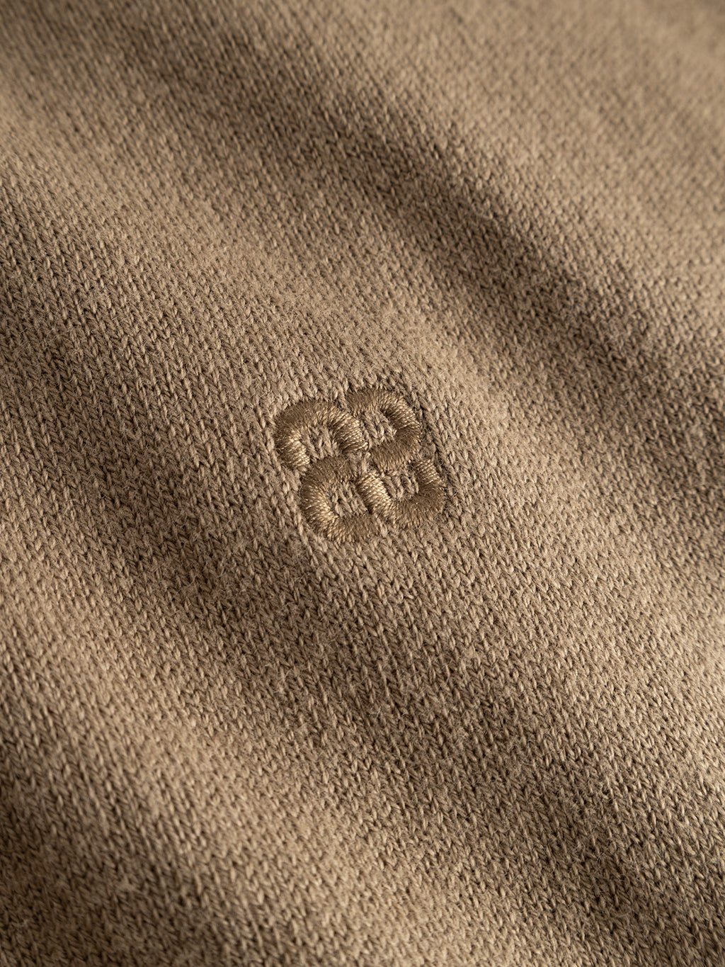 Close-up of light brown fabric from the Gabba GABfumo Element Spy LS Knit - Sand Crocker sweater, with a subtle embroidered geometric logo in the center featuring four interlocking shapes forming a square pattern. The texture of this crew neck knit is soft and slightly ribbed.