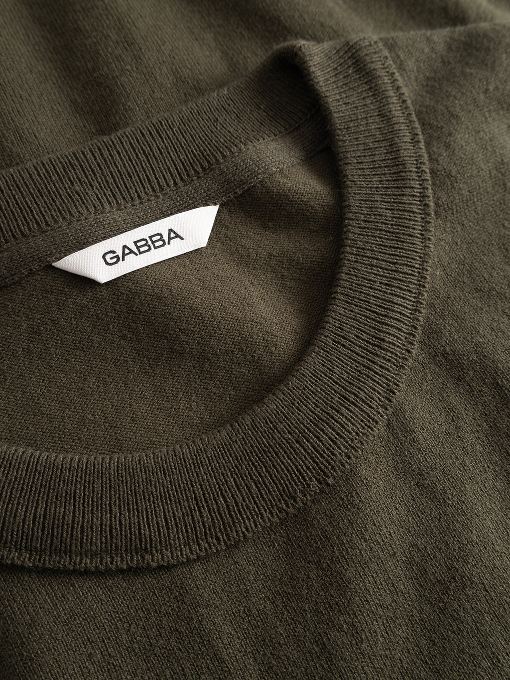 Close-up of the GABfumo Element Spy LS Knit in Forest Night, an olive green crewneck sweater made from flat-knit cotton, featuring a white tag with the Gabba brand name in black text inside the collar.