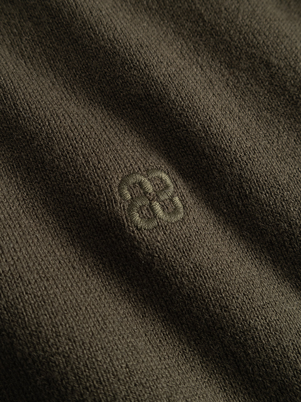 A close-up of the GABfumo Element Spy LS Knit in Forest Night by Gabba reveals its textured, dark olive flat-knit cotton weave, with a small embroidered geometric logo at the center. The detailed stitching and premium material quality are clearly visible.