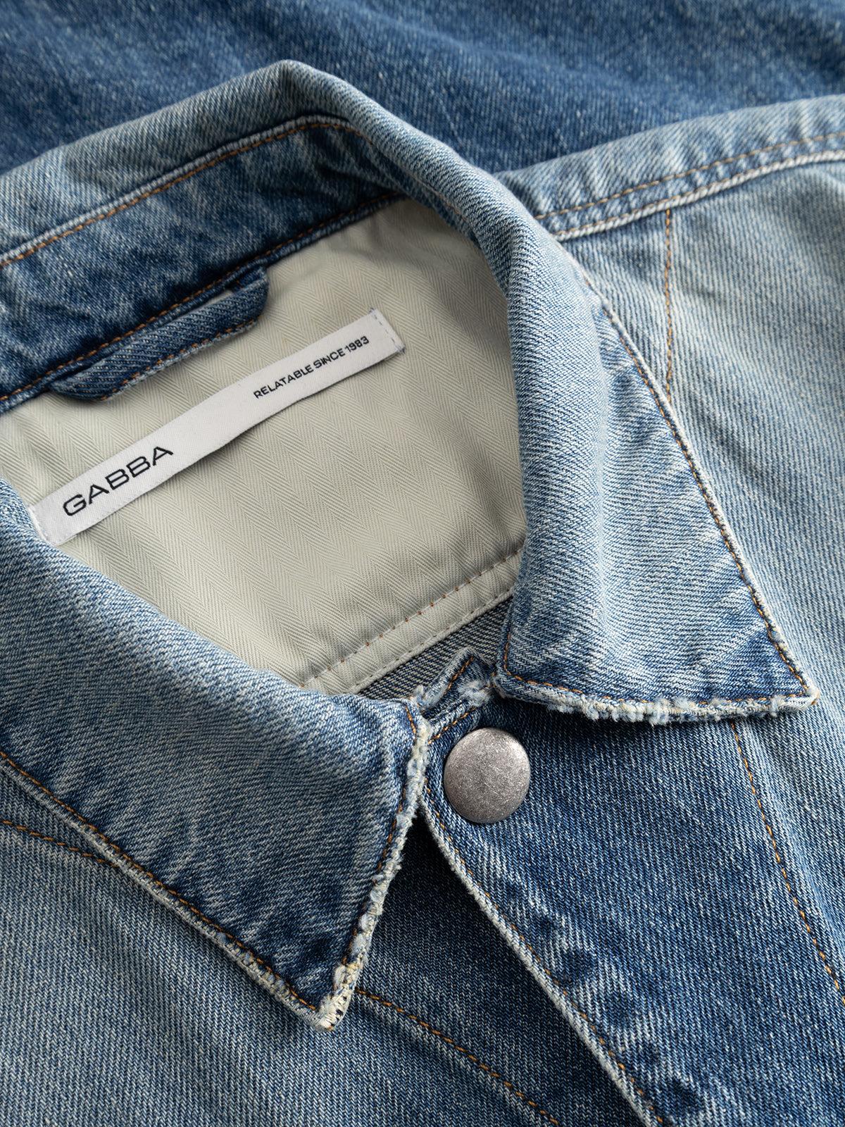 Close-up of the GABbeta Wolv DS0004 LS Overshirt in dark blue denim by Gabba, featuring a metal button and detailed collar with an inner label that reads Gabba. The workwear-inspired design emphasizes the rich fabric texture and precise stitching, while a laser-washed finish accentuates its distinctive character.