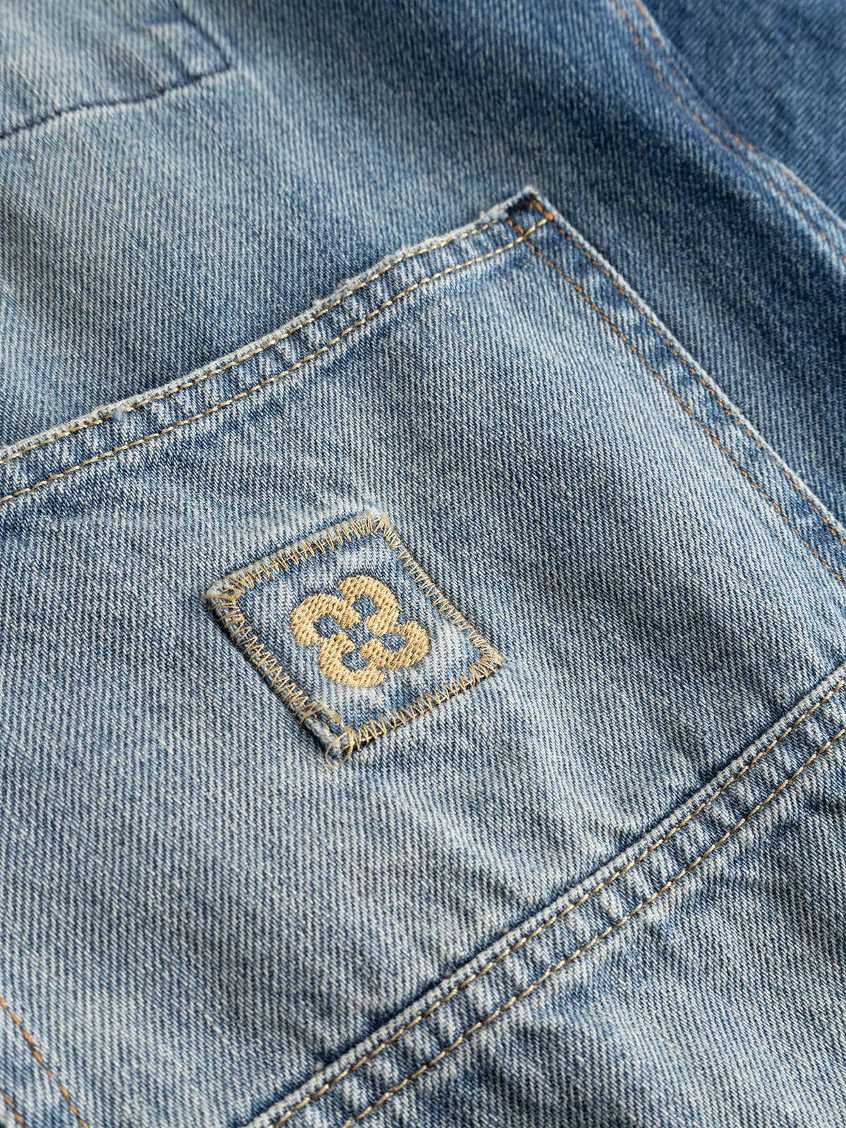 Close-up of the Gabba GABbeta Wolv DS0004 LS Overshirt in dark blue denim, highlighting its workwear-inspired design with a front pocket. A small stitched square patch displays a yellow embroidered motif resembling a stylized flower or geometric pattern, all set against the distinctive laser-washed cotton fabric.