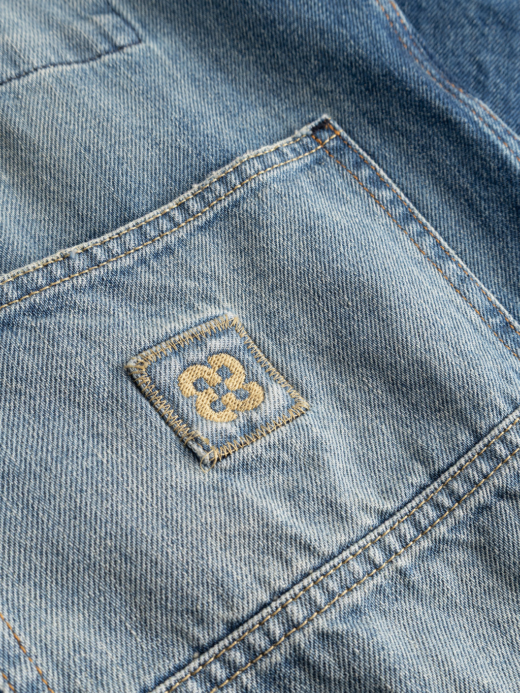 Close-up of the Gabba GABbeta Wolv DS0004 LS Overshirt in dark blue denim, highlighting its workwear-inspired design with a front pocket. A small stitched square patch displays a yellow embroidered motif resembling a stylized flower or geometric pattern, all set against the distinctive laser-washed cotton fabric.