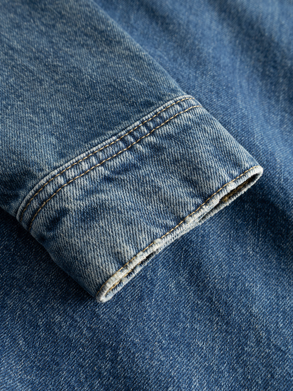 A close-up of the sleeve of the Gabba GABbeta Wolv DS0004 LS Overshirt in dark blue denim, displayed on matching blue denim fabric. The visible stitching along the cuff accentuates its laser-washed finish, emphasizing its workwear-inspired design.