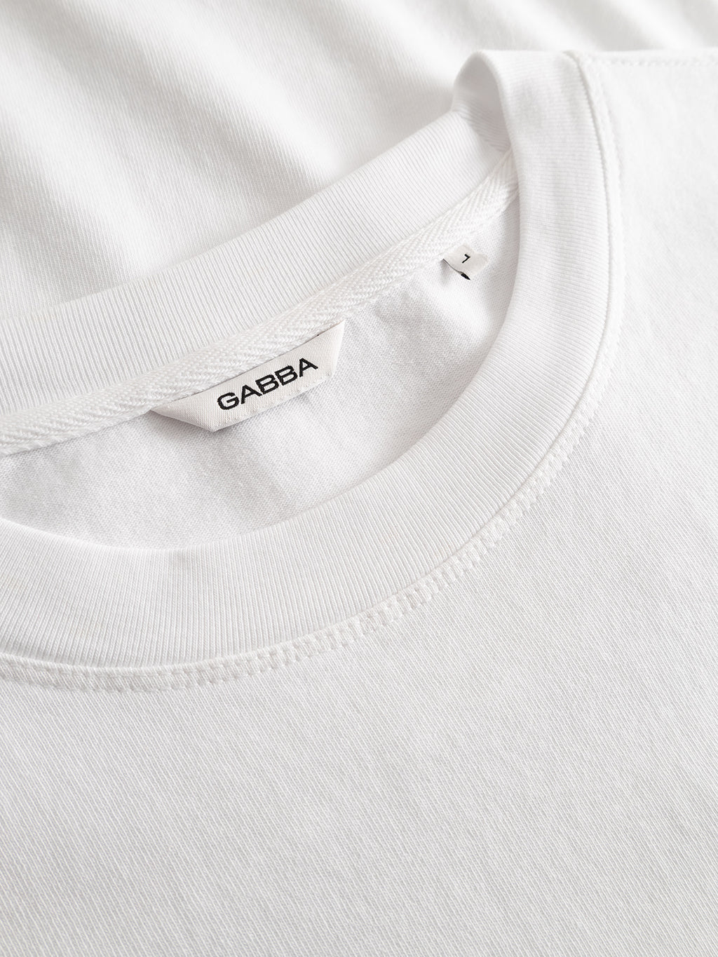 Close-up of the collar of the Gabba GABlegin Wix Chap Boxy Hour SS T-Shirt in white, featuring a Gabba brand label and size L tag, laid flat on a matching white surface.