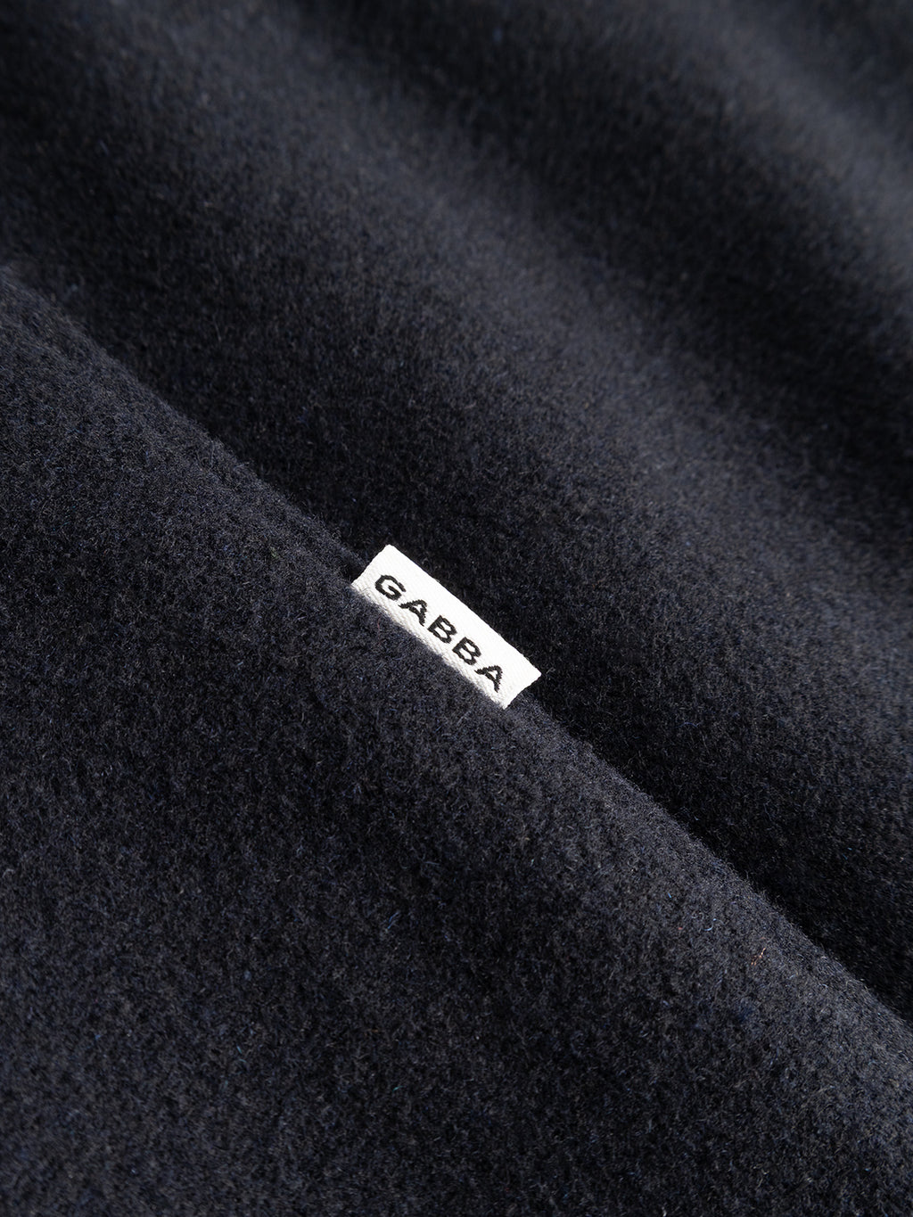 A close-up of the GABleeds Plain Coat in navy by Gabba showcases its dark, textured fabric and a small white label bearing the brand’s name in bold black capitals—an understated detail that embodies the elevated craftsmanship of this timeless outerwear piece.