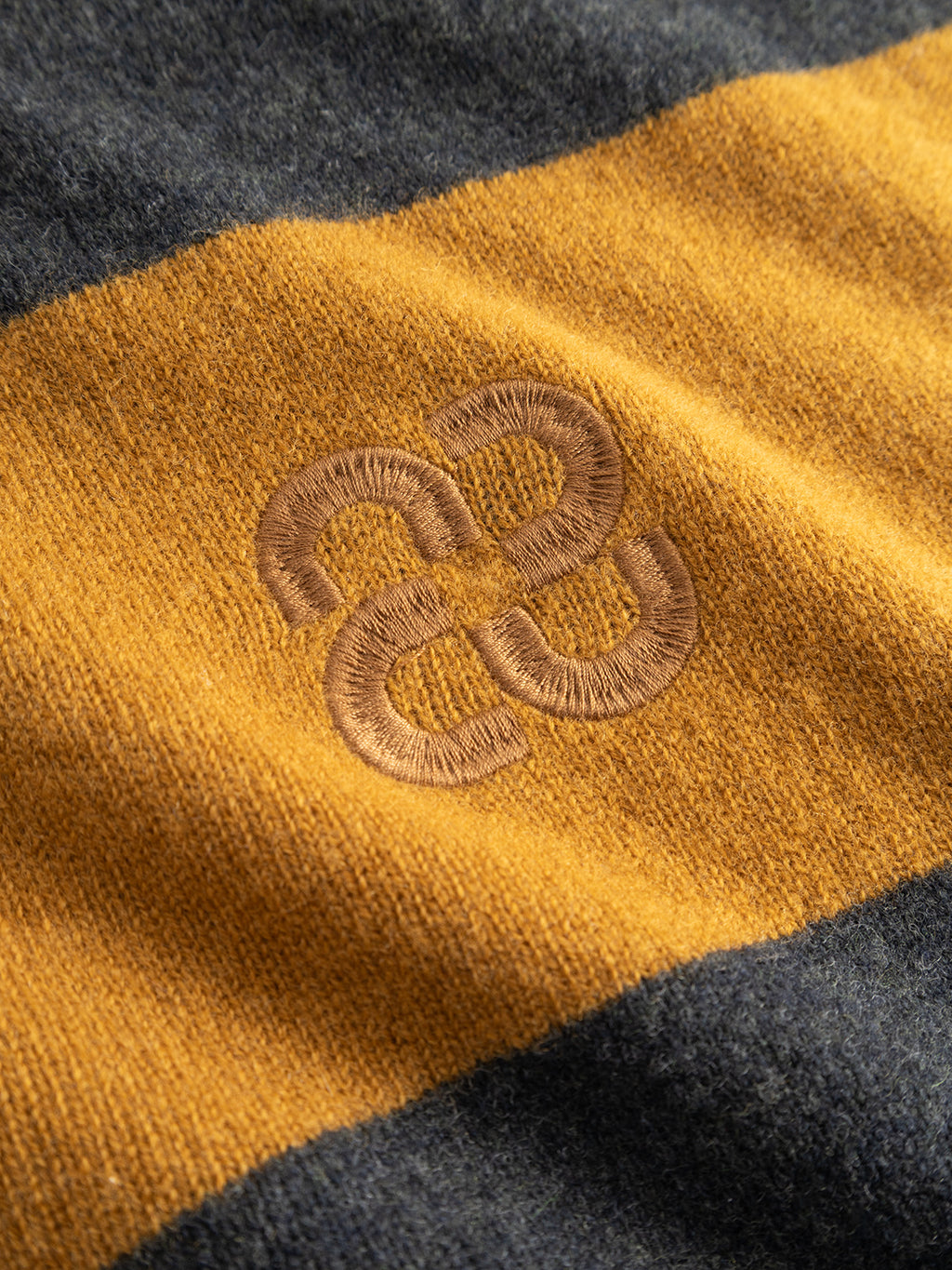 A close-up of the Gabba GABwest Aico Scarf - Yellow Stripes showcases its merino wool fabric with a wide mustard yellow stripe bordered by dark gray, and a stitched brown geometric logo centered on the yellow section. This fashionable winter accessory from Gabba offers exceptional warmth, comfort, and distinctive style.