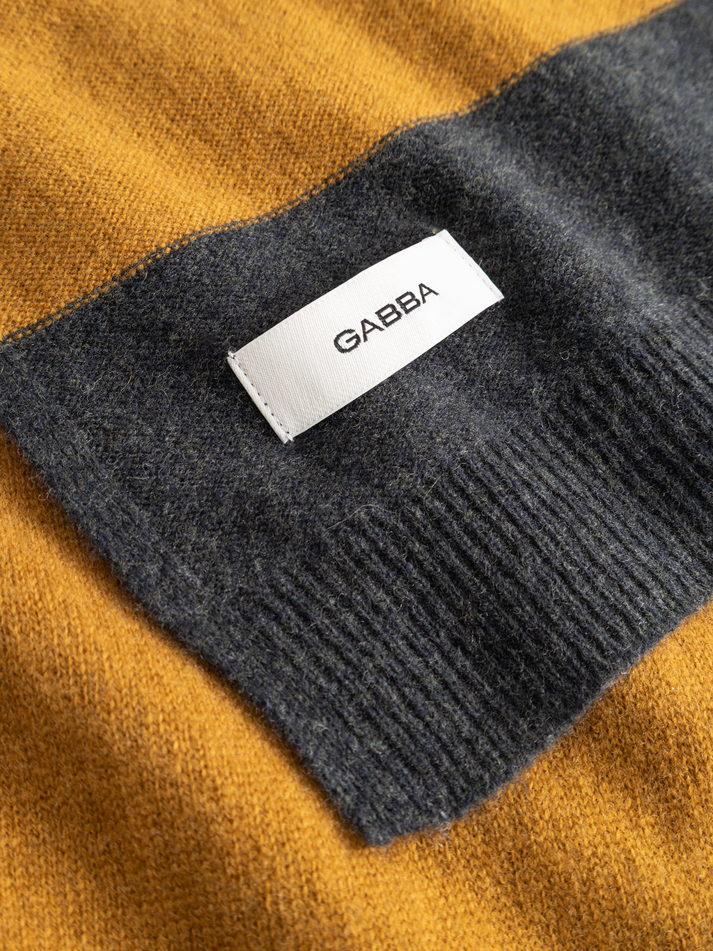 A close-up image shows a sweater with mustard yellow and dark gray knit fabric, highlighted by a white “GABBA” label stitched on the gray section, next to the GABwest Aico Scarf - Yellow Stripes from Gabba—a lightweight accessory crafted from soft merino wool, perfect for winter.