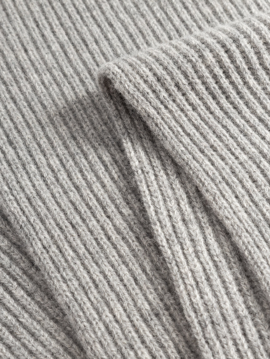 Close-up of the GABeast Flash Scarf - Grey Mel by Gabba, showcasing its light gray knitted fabric with a ribbed texture. The image highlights the detailed fibers and a folded section that emphasizes the thickness and softness of the scarf, making it an ideal winter accessory.
