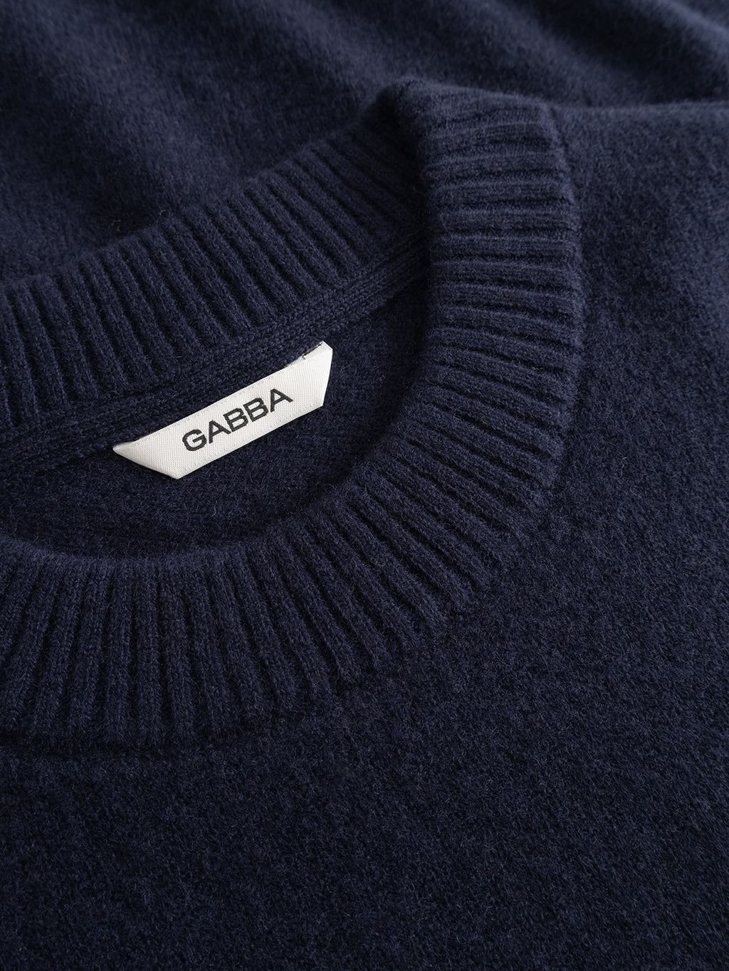 Close-up of the GABtokota Element Aico LS Knit in navy, featuring a crew neck with a ribbed neckline. A white tag inside the collar displays the Gabba brand name in black letters. The soft wool knit sweater offers a textured feel for everyday comfort.