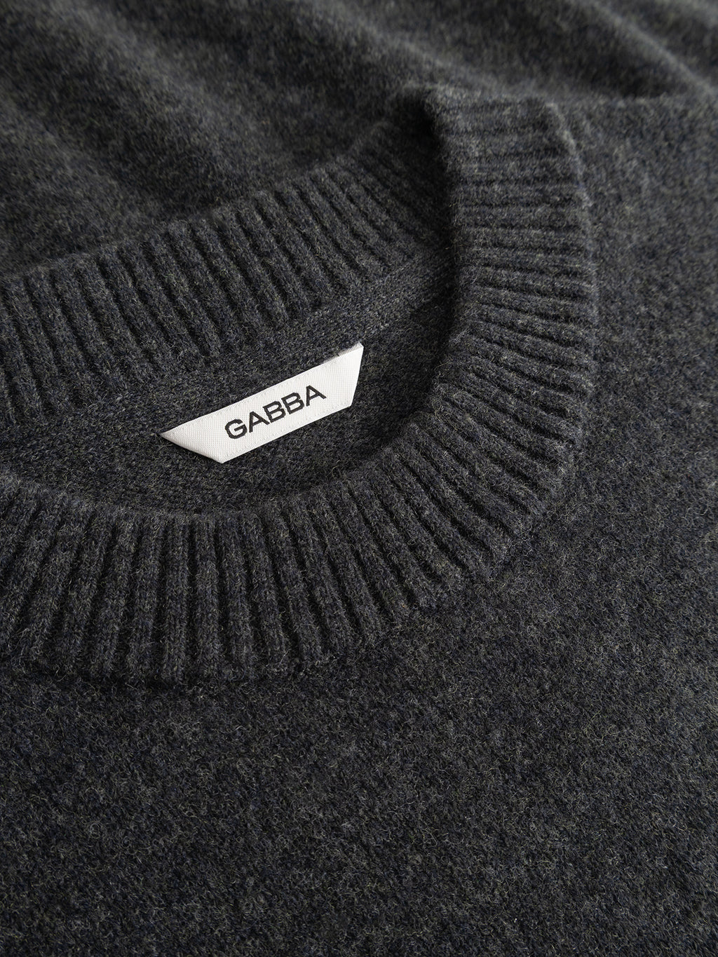 Close-up of the GABtokota Element Aico LS Knit in Dark Grey Mel. by Gabba, a regular fit knitted sweater featuring a ribbed crew neckline. A white label inside the collar reads Gabba in black letters, emphasizing the timeless design of this soft wool sweater.