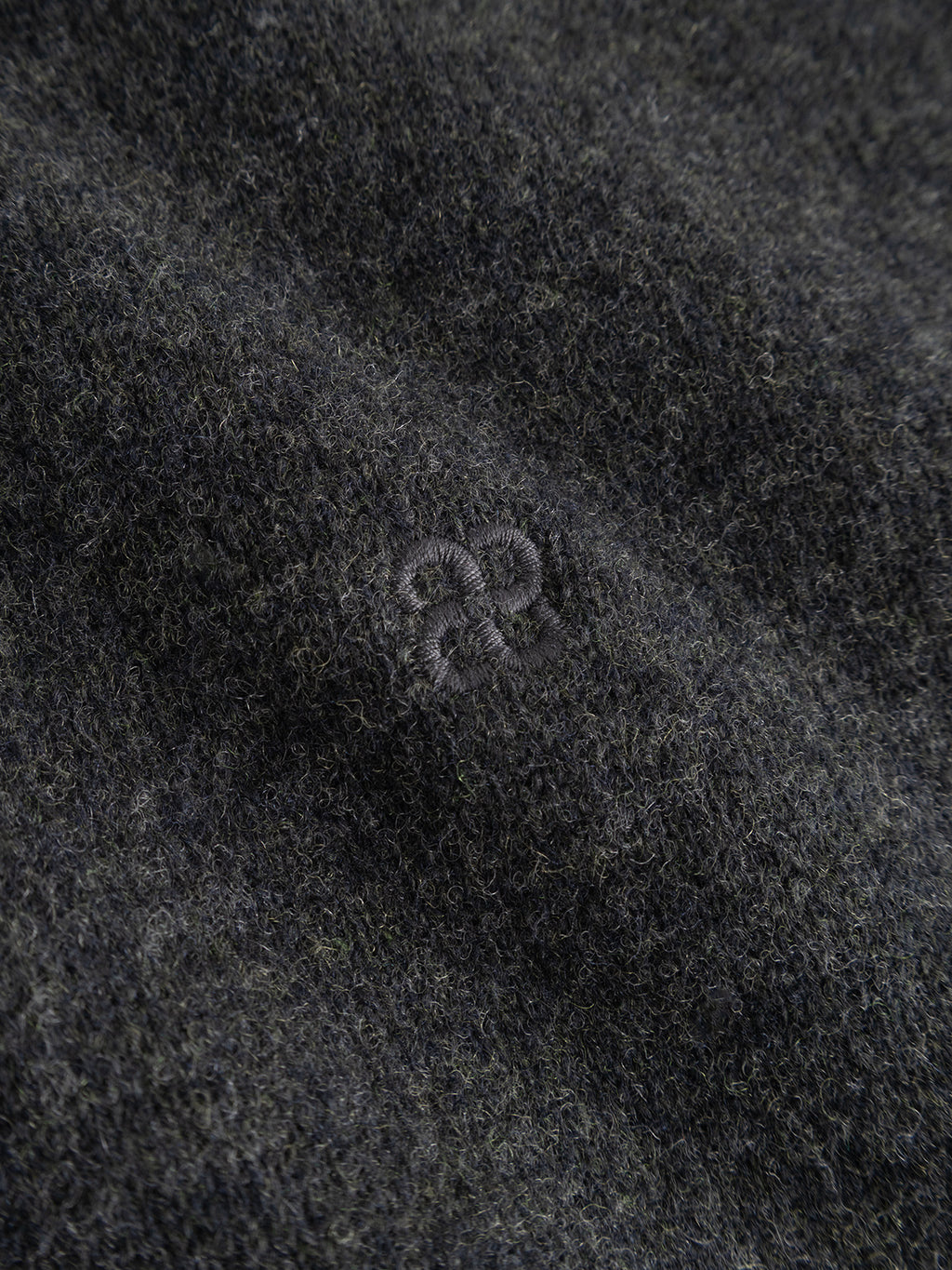 A close-up of the GABtokota Element Aico LS Knit in Dark Grey Mel. by Gabba reveals its dark gray, textured wool fabric with subtle stitching forming a small, circular embroidered pattern near the center—perfectly capturing the refined details and regular fit of this soft sweater.
