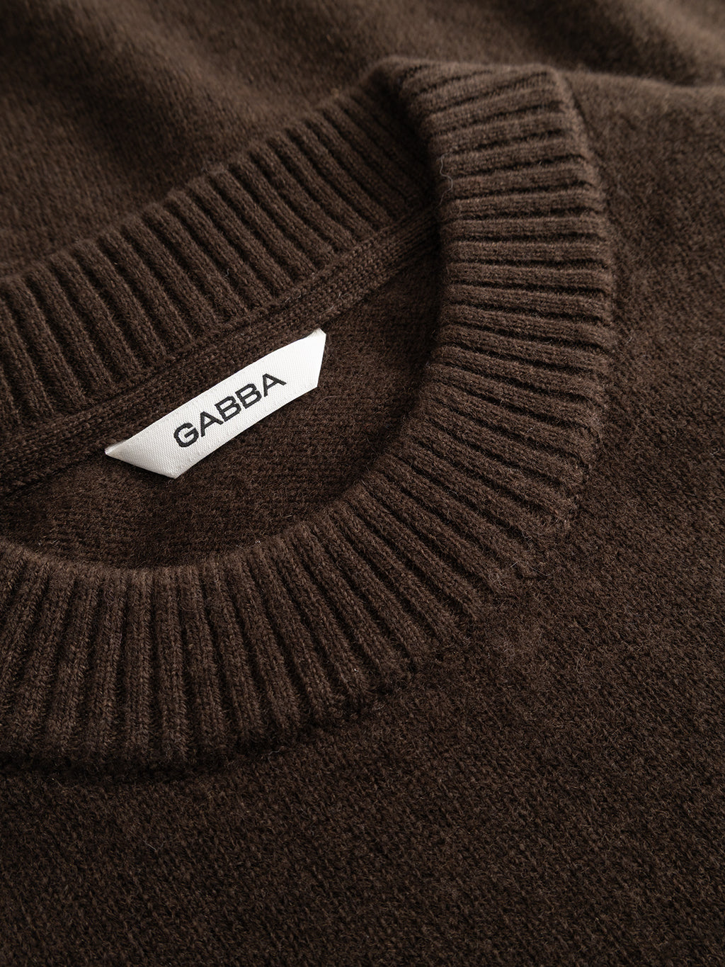 Close-up of the GABtokota Element Aico LS Knit sweater by Gabba in brown slate, featuring a ribbed crew neck and a white tag with the brand name.