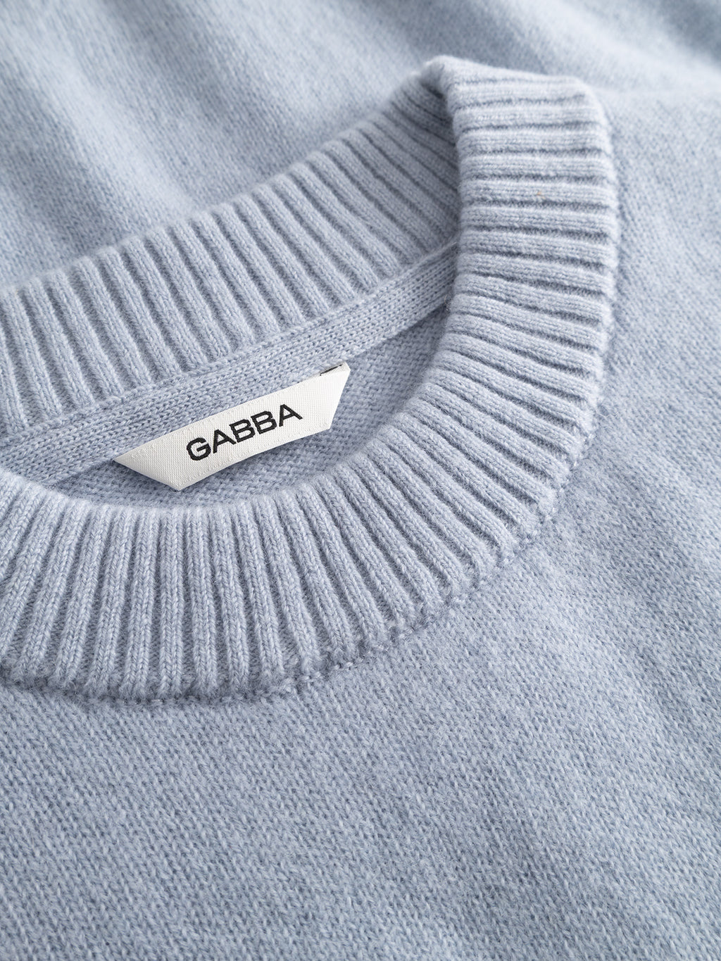 The GABtokota Element Aico LS Knit by Gabba is a dusty blue crew neck sweater featuring a ribbed collar and crafted for everyday comfort. A white tag inside the collar displays the brand name Gabba in bold black uppercase letters. Made from soft wool, this knit offers a cozy texture, making it perfect for versatile wear.