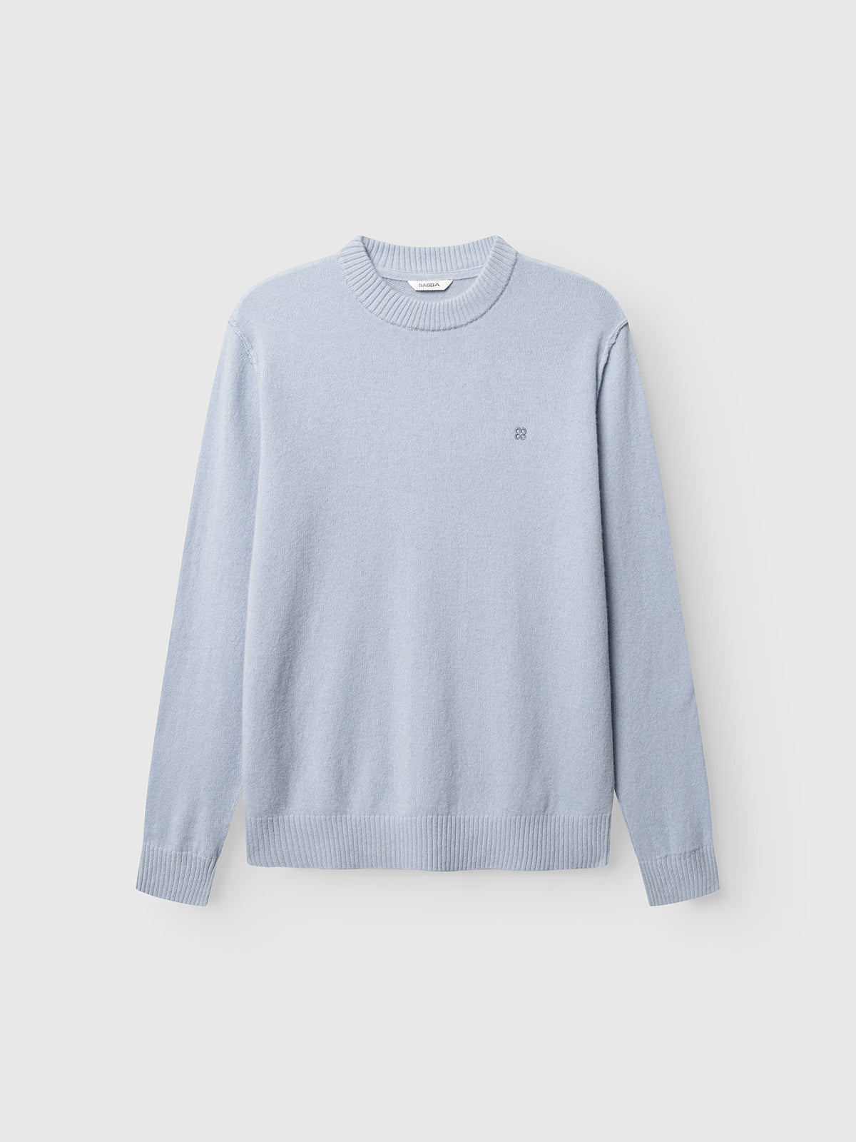 The GABtokota Element Aico LS Knit in Dusty Blue by Gabba is a light blue, long-sleeve crewneck sweater featuring ribbed cuffs, hem, and neckline for everyday comfort. A small, subtle Gabba logo is embroidered on the left chest. This soft wool sweater is shown laid flat against a plain white background.