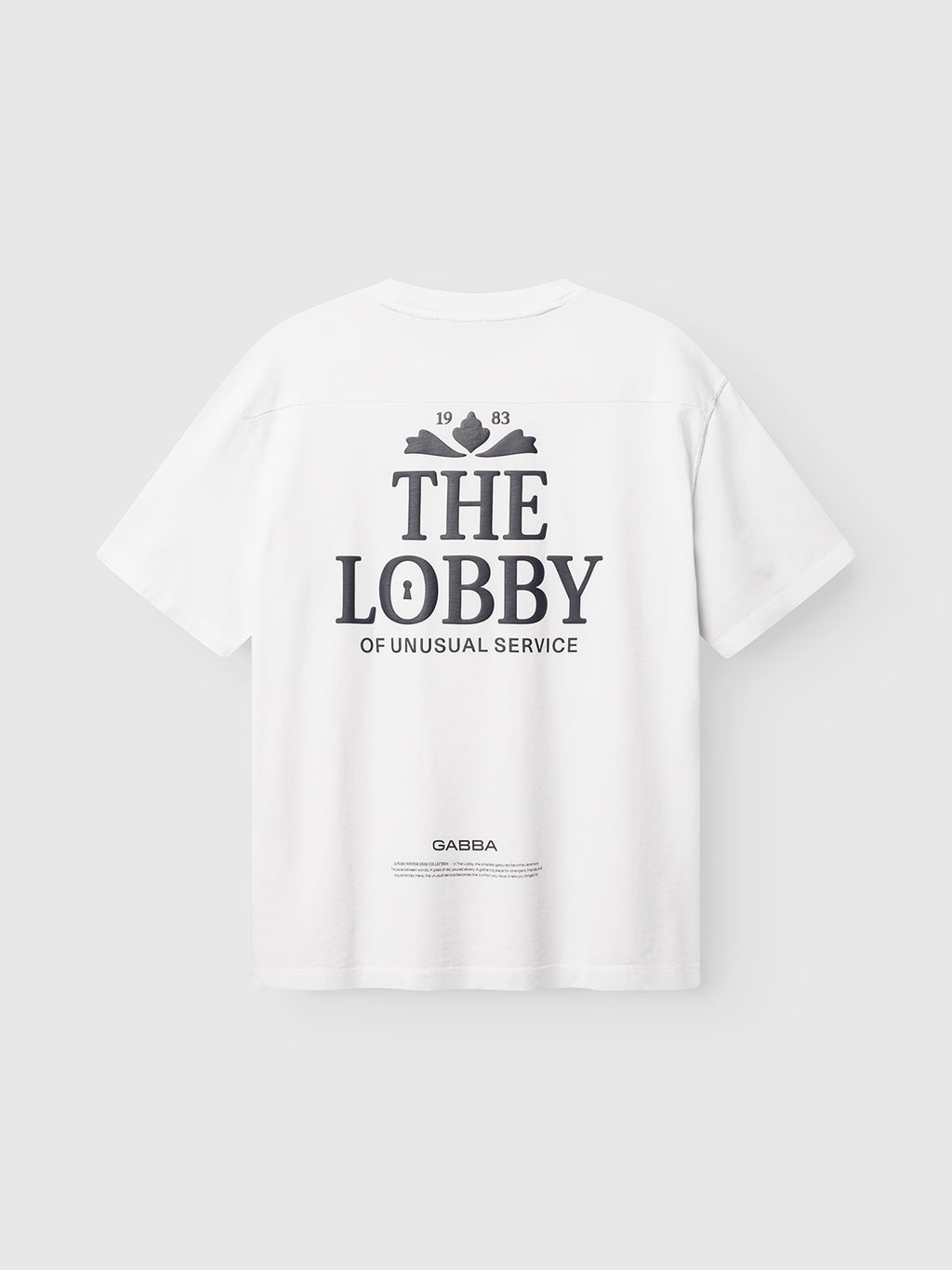 The GABlegin Wix Boxy Hour Lobby1 SS T-shirt by Gabba is a white, relaxed boxy-fit tee crafted from textured cotton jersey. It features bold black “THE LOBBY OF UNUSUAL SERVICE” and “GABBA” text printed on the back, complemented by a small illustration of two crossed keys and the year 1983.