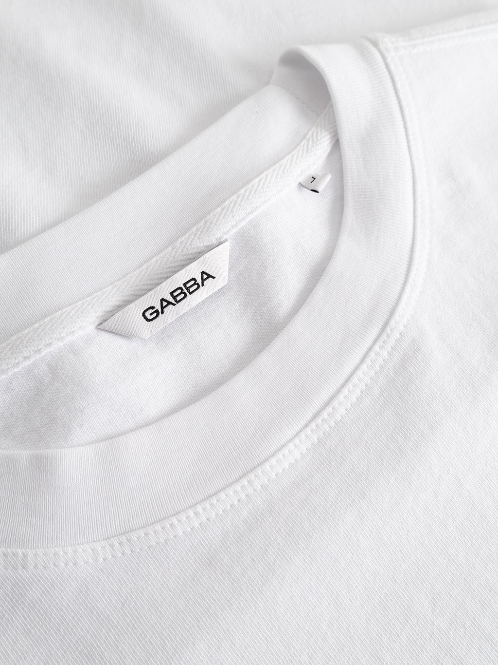 Close-up of the Gabba GABlegin Wix Boxy Hour Lobby1 SS T-shirt in white, featuring a visible Gabba label sewn into the neckline. Made from textured cotton jersey, the T-shirt appears new and is neatly folded on a white surface.