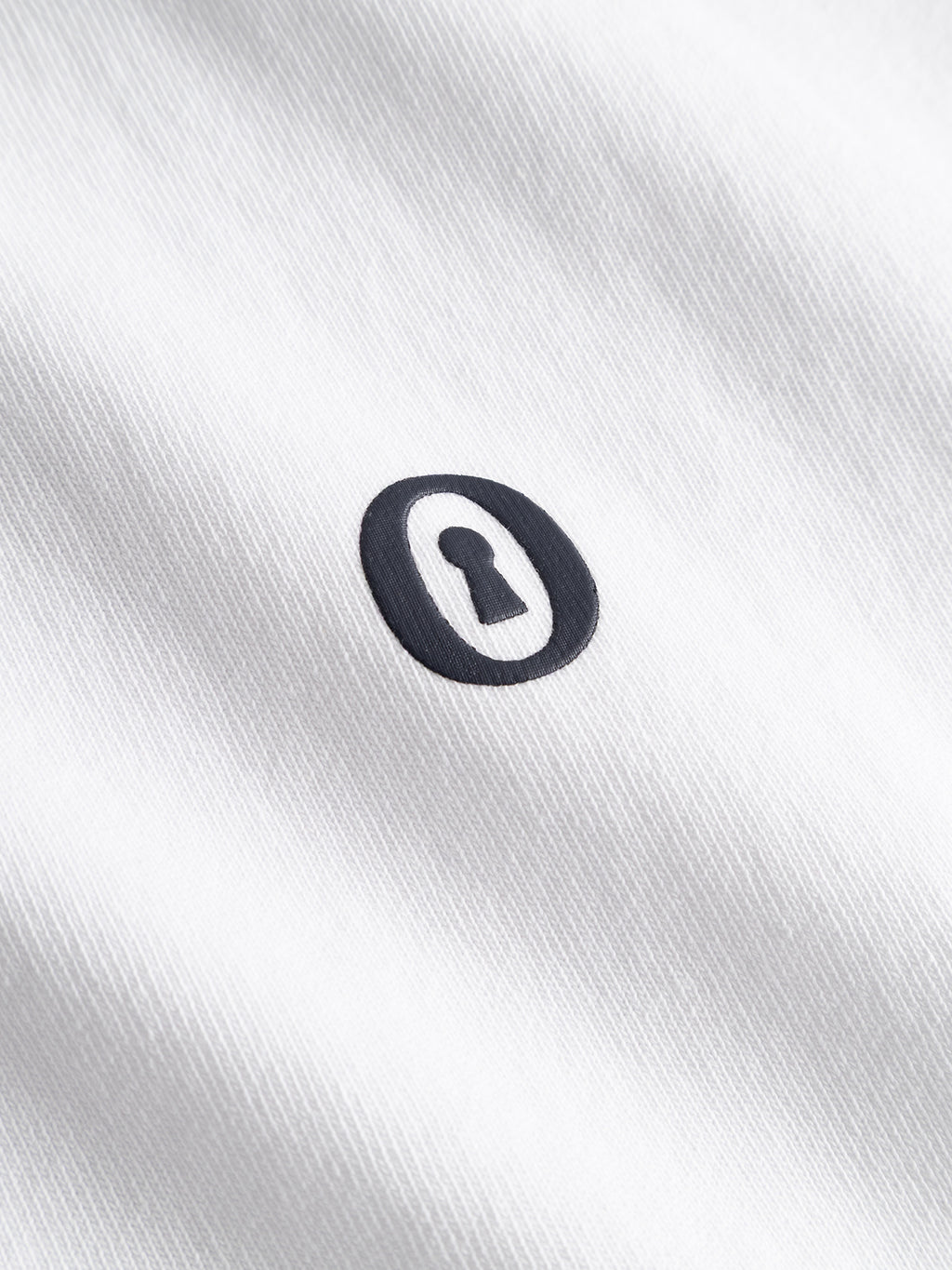 A black keyhole symbol inside a black oval is printed on the white, textured cotton jersey fabric of the Gabba GABlegin Wix Boxy Hour Lobby1 SS T-shirt.
