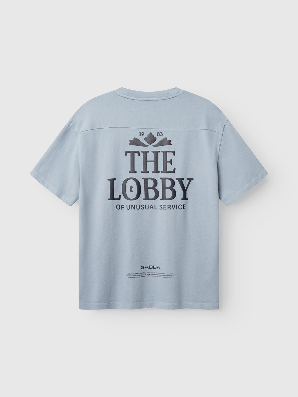 Introducing the Gabba GABlegin Wix Boxy Hour Lobby1 SS T-shirt in Dusty Blue—crafted from textured cotton jersey with a relaxed, boxy fit. The back features a bold graphic print displaying “1983 THE LOBBY OF UNUSUAL SERVICE” in large black letters, accompanied by additional smaller text below.