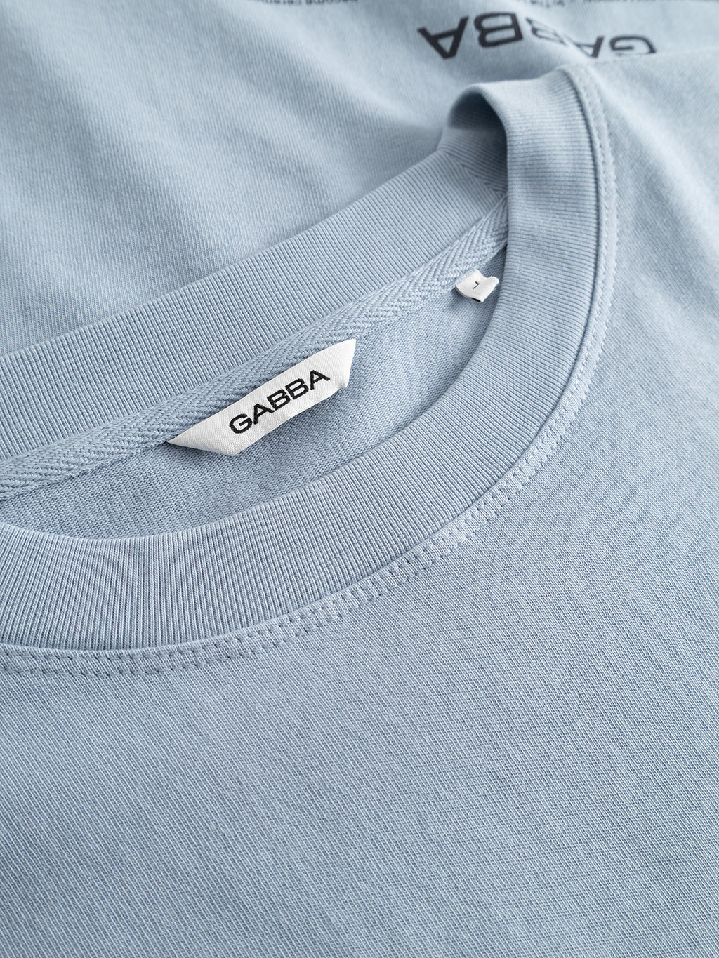 Close-up of the GABlegin Wix Boxy Hour Lobby1 SS T-shirt in Dusty Blue by Gabba, featuring a white brand tag. Made from textured cotton jersey, this crewneck style offers a relaxed, boxy fit. A glimpse of another similarly colored shirt appears in the background.
