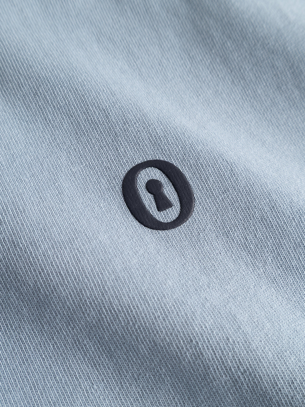Close-up of the dusty blue textured cotton jersey fabric of the Gabba GABlegin Wix Boxy Hour Lobby1 SS T-shirt, featuring a black oval logo with a keyhole design at the center. The visible fabric texture highlights the relaxed, boxy fit.