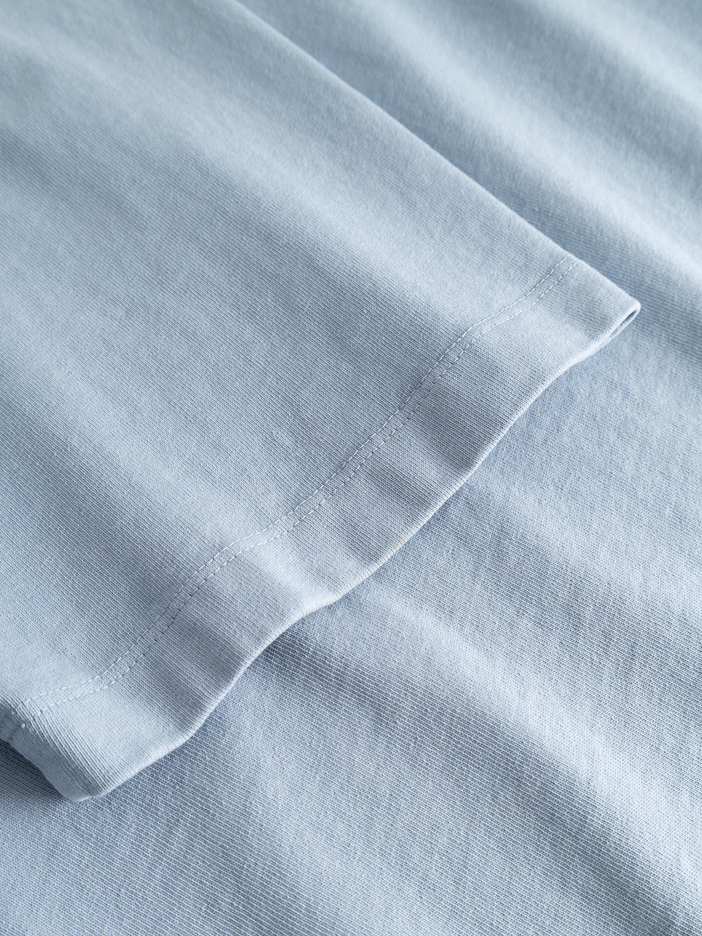 A close-up of the hem of the Gabba GABlegin Wix Boxy Hour Lobby1 SS T-shirt in Dusty Blue, lying flat on matching blue fabric. The stitching and texture of the cotton jersey material are clearly visible.