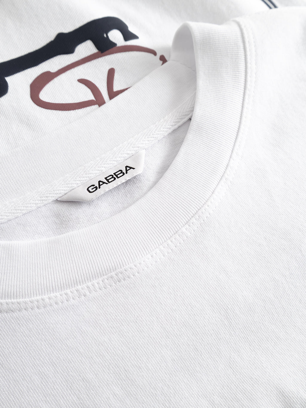 Close-up of the Gabba GABlegin Wix Boxy Hour Lobby2 SS T-shirt in white, crafted from textured cotton jersey with a visible Gabba label at the collar and a partial printed graphic in black and brown on the fabric.