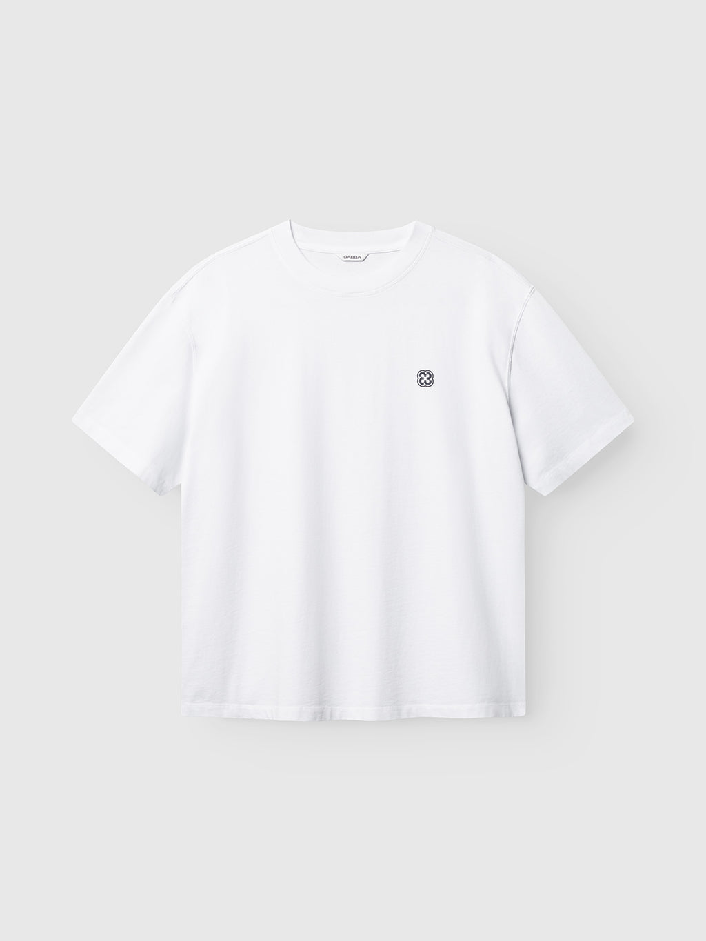 Introducing the Gabba GABlegin Wix Boxy Hour Lobby2 SS T-shirt in white—a short-sleeve tee featuring a small, circular black logo on the left chest. Crafted from textured cotton jersey, this relaxed boxy-fit t-shirt delivers everyday comfort and modern style, shown here against a light gray background.