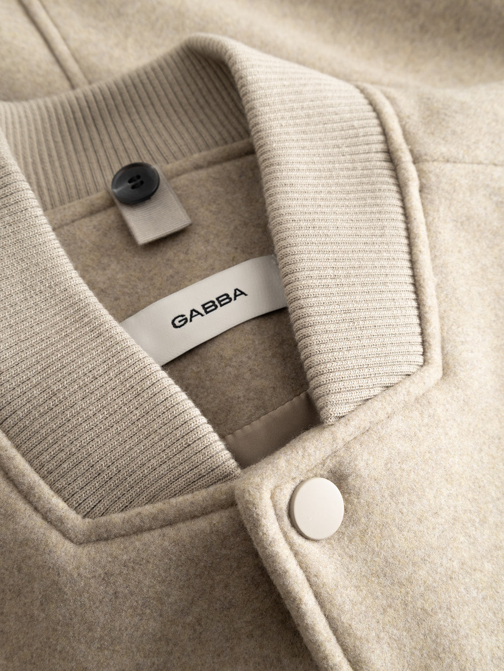 Close-up of the GABmatheo Plain Bomber Jacket in Sand by Gabba, featuring a beige wool-inspired fabric, ribbed collar, snap button closure, and an inner label with the brand name—an ideal piece of casual outerwear.