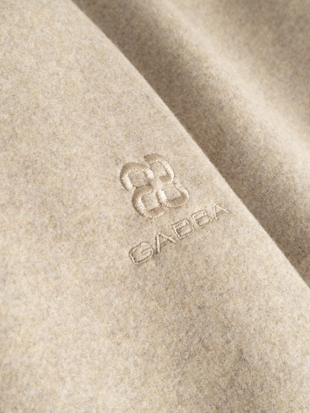 Close-up of the soft, light beige fabric of the Gabba GABmatheo Plain Bomber Jacket in Sand, featuring a geometric embroidered logo with “GABBA” beneath it. The wool-inspired material appears cozy and ideal for casual outerwear.