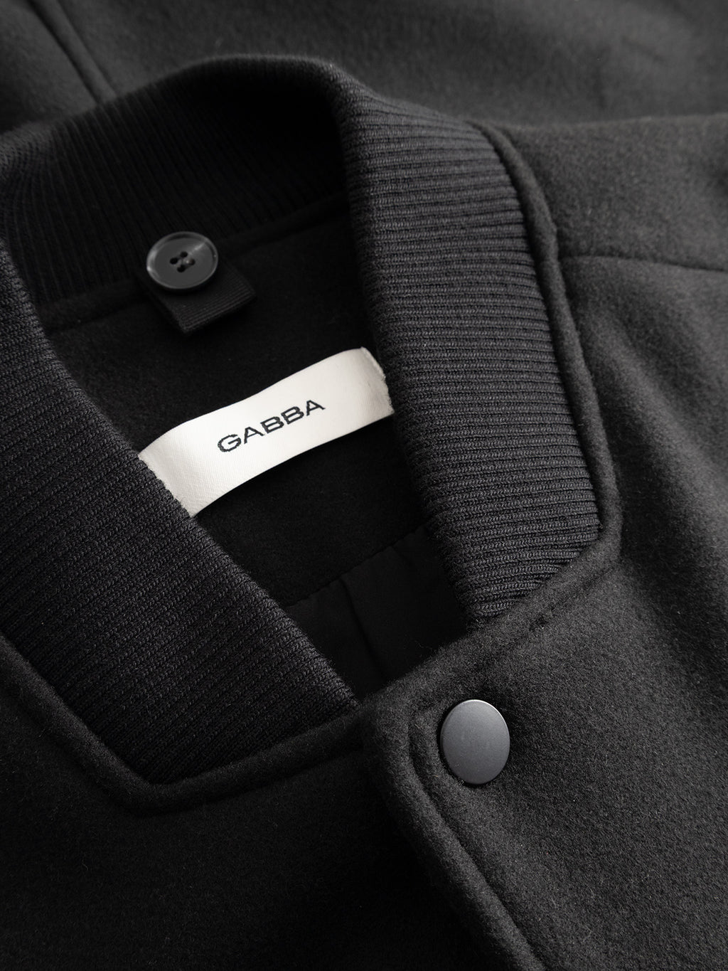 Close-up of the Gabba GABmatheo Plain Bomber Jacket in black, featuring a ribbed collar, button details, and a white inner label with the Gabba logo. Made from wool-inspired fabric, this jacket is an ideal option for casual outerwear.