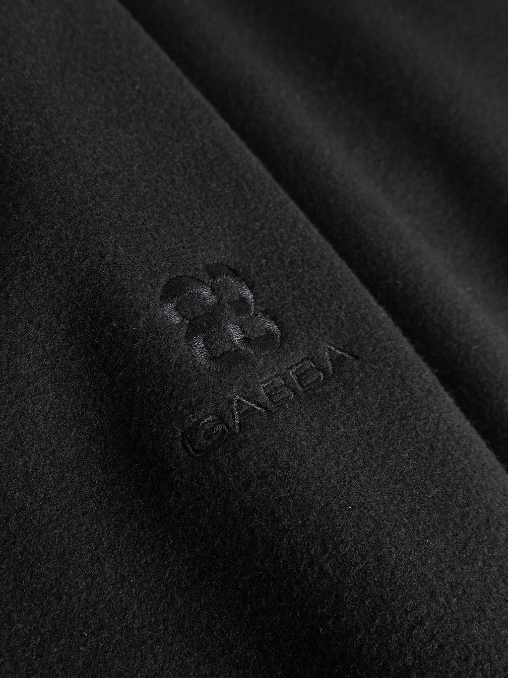 Close-up of the GABmatheo Plain Bomber Jacket by Gabba, featuring black wool-inspired fabric with a soft, subtly fuzzy texture reminiscent of premium casual outerwear. Above the word “GABBA,” a subtle embroidered flower-like logo blends in with the black stitching and material.