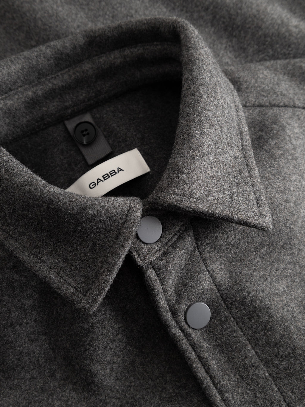 A close-up of the GABtheo City Jacket in dark grey, folded to showcase its snap buttons and the visible Gabba brand tag on the collar. Crafted from a soft, wool-inspired fabric, this jacket is perfect for everyday layering.