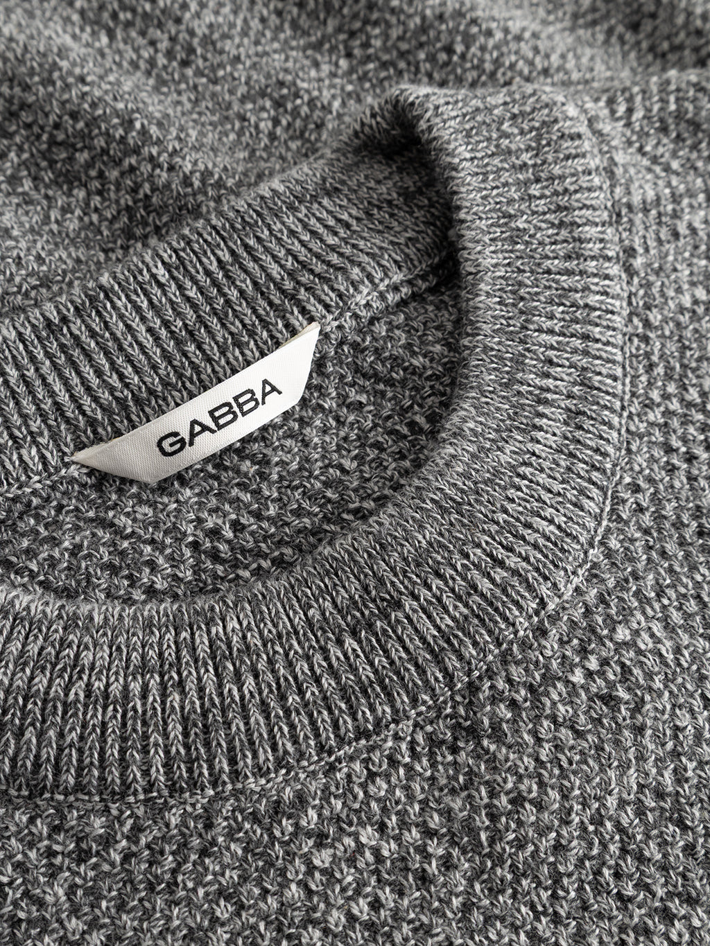 Close-up of the GABlamix Coro O-Neck LS Knit in light grey, a textured cotton-blend sweater with a round neckline. A white fabric tag with the Gabba brand name in black letters is visible near the collar.