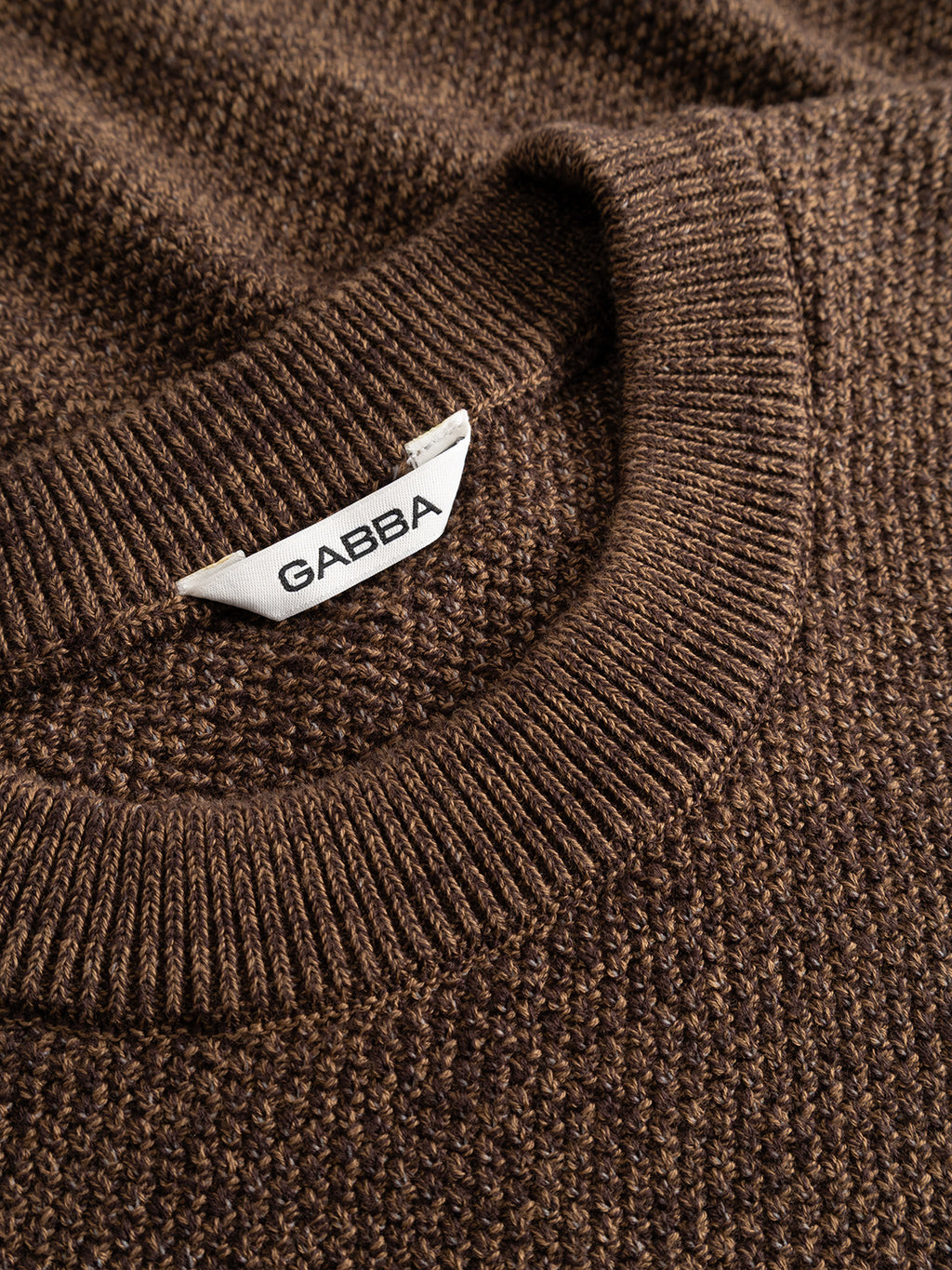 Close-up of the GABlamix Coro O-Neck LS Knit in Brown Slate by Gabba, featuring a brown cotton-blend knit fabric, a ribbed crew neck, and a white Gabba tag sewn onto the inside collar.
