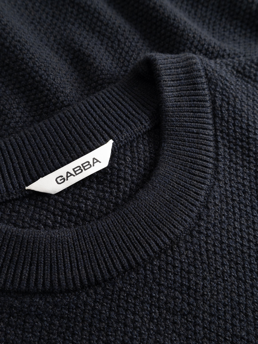Close-up of the GABlamix Coro O-Neck LS Knit sweater in blue black, a dark textured cotton-blend knit with a ribbed crew neck collar. This regular fit sweater features a white fabric tag on the collar displaying the Gabba brand name in black capital letters.